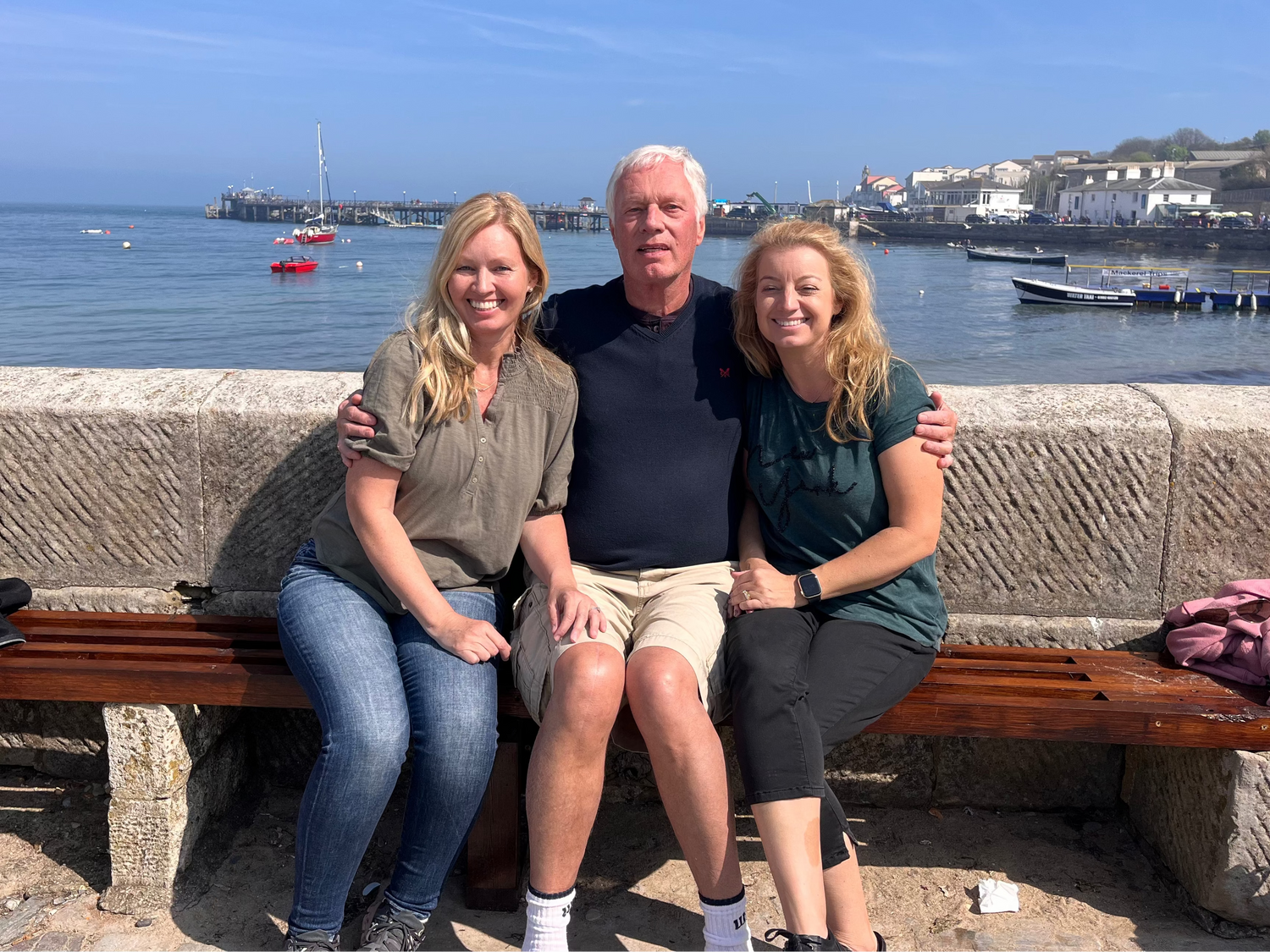 Three people sitting on a bench by a waterfront with boats and buildings in the background.